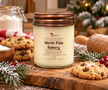 A North Country Candle jar candle labeled 'North Pole Bakery' sits on a wooden surface surrounded by cookies, evergreen branches, red berries, and a warm holiday background.